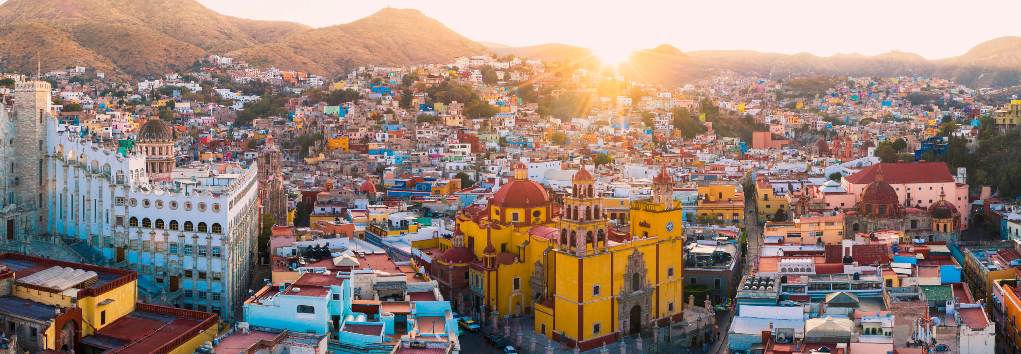 Vibrant city of Mexico, showcasing its colorful buildings and mountains in distance, the sun setting beyond