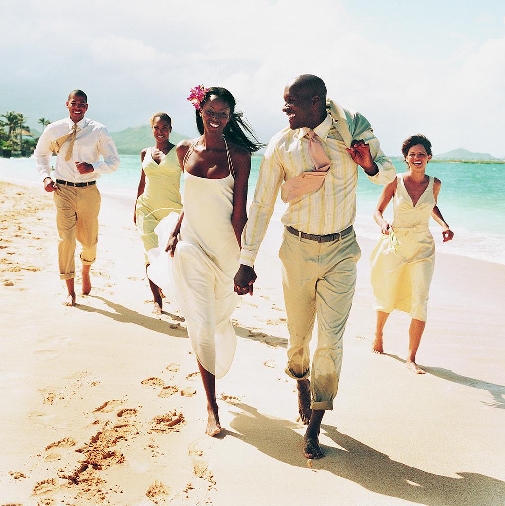 A newlywed couple walk along a beach with their wedding party close behind