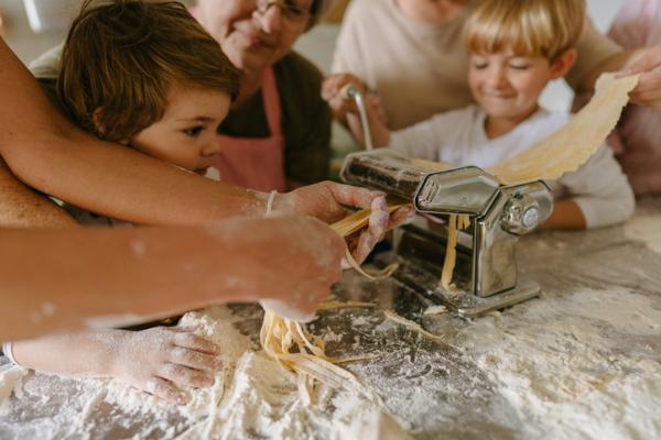 Children using a pasta machine with adults helping in a flour-covered kitchen