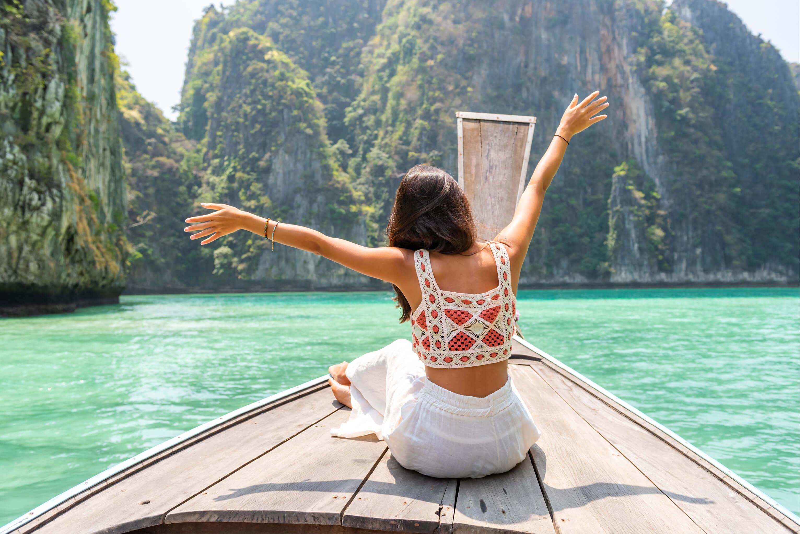 Person on a boat joyfully extending her arms wide against the backdrop of turquoise water, lush mountains, and sky