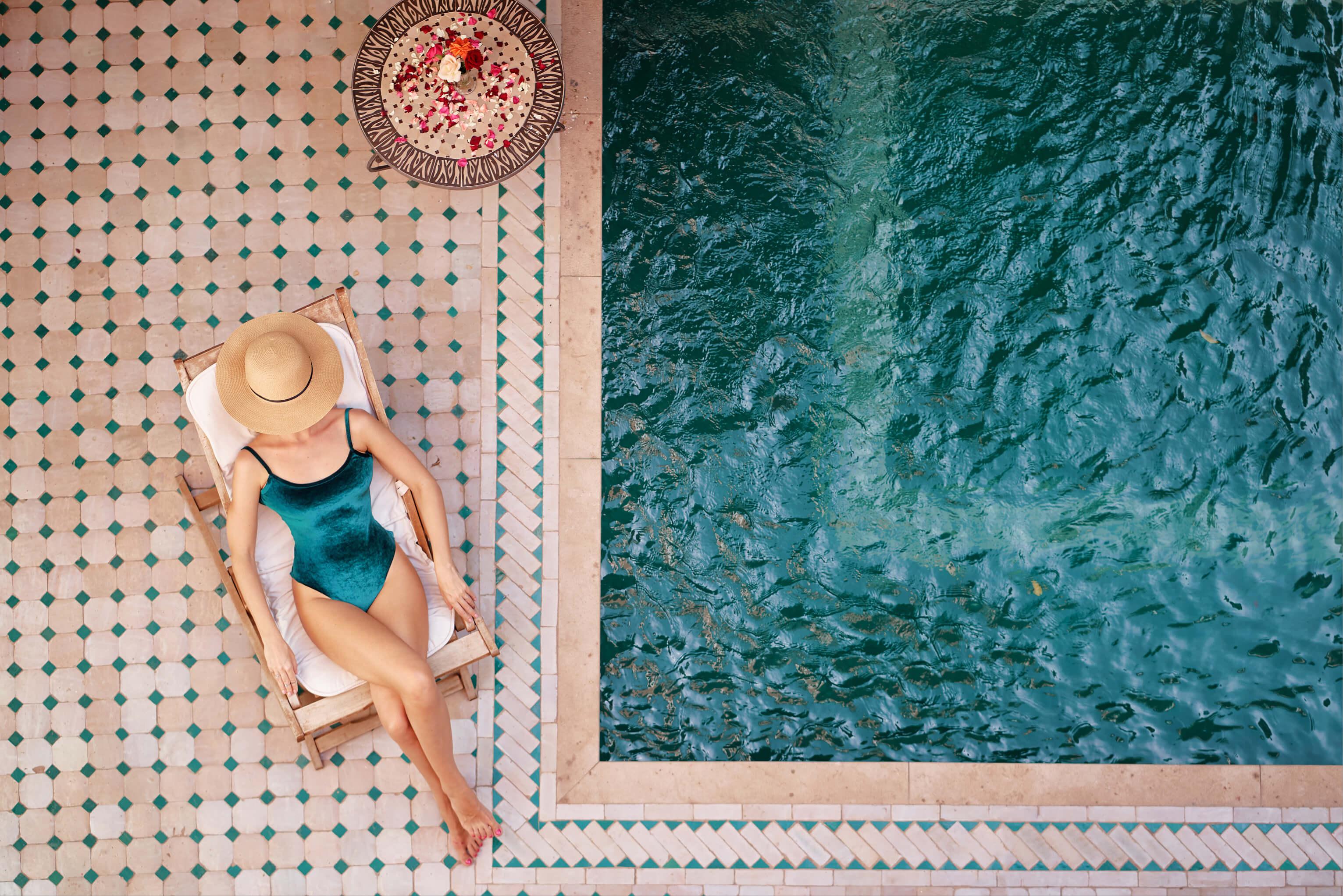 A woman in a swimsuit with a hat covering her face relaxes in a chair beside a sparkling pool on a sunny day