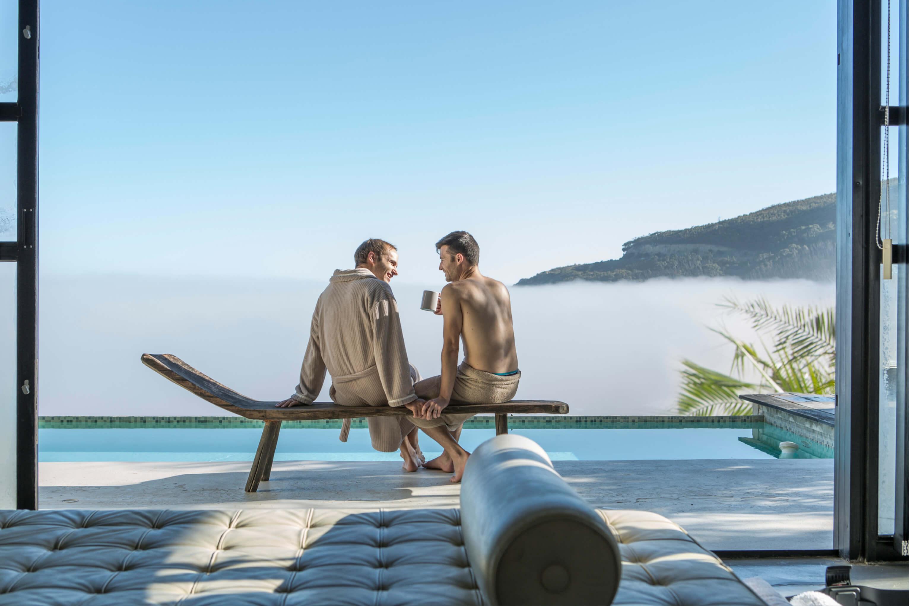 Two men sit on a lounge chair near a pool, engaged in conversation amidst a serene outdoor environment