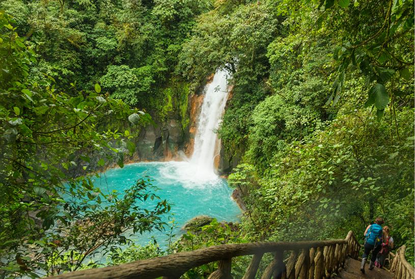Travelers descend wooden stairs through the rainforest in Central America, heading towards a cascading waterfall
