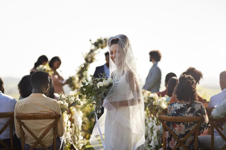 Smiling bride in veil holding flowers at outdoor wedding with guests seated