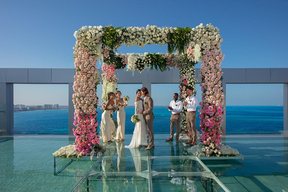 Rooftop wedding with a glass floor, featuring a pink and white floral arch, bride and groom, bridesmaids in white dresses, and groomsmen in tan suits, overlooking the ocean.